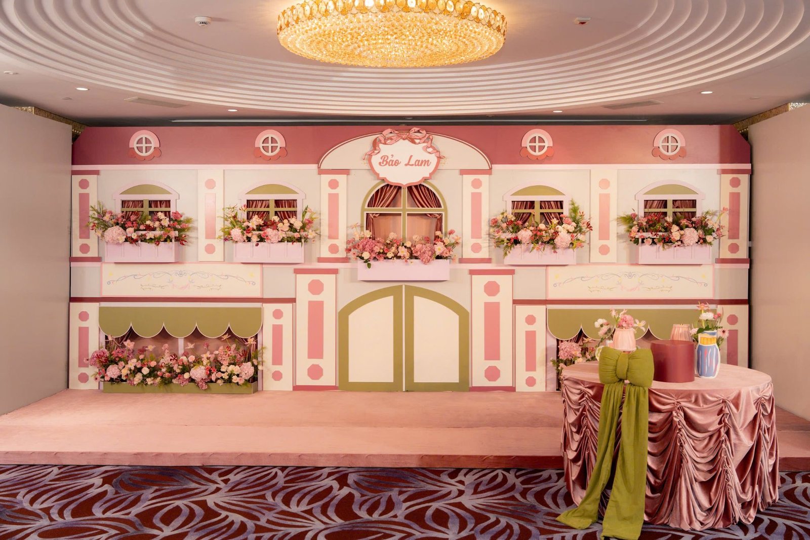 Backdrop ngôi nhà hồng thực tế với bảng tên Bảo Lam và bàn bánh phủ nhung
