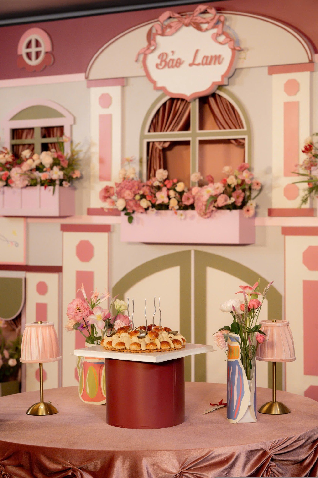 Bàn bánh sinh nhật với đèn bàn hồng và bình hoa vẽ tay trước backdrop