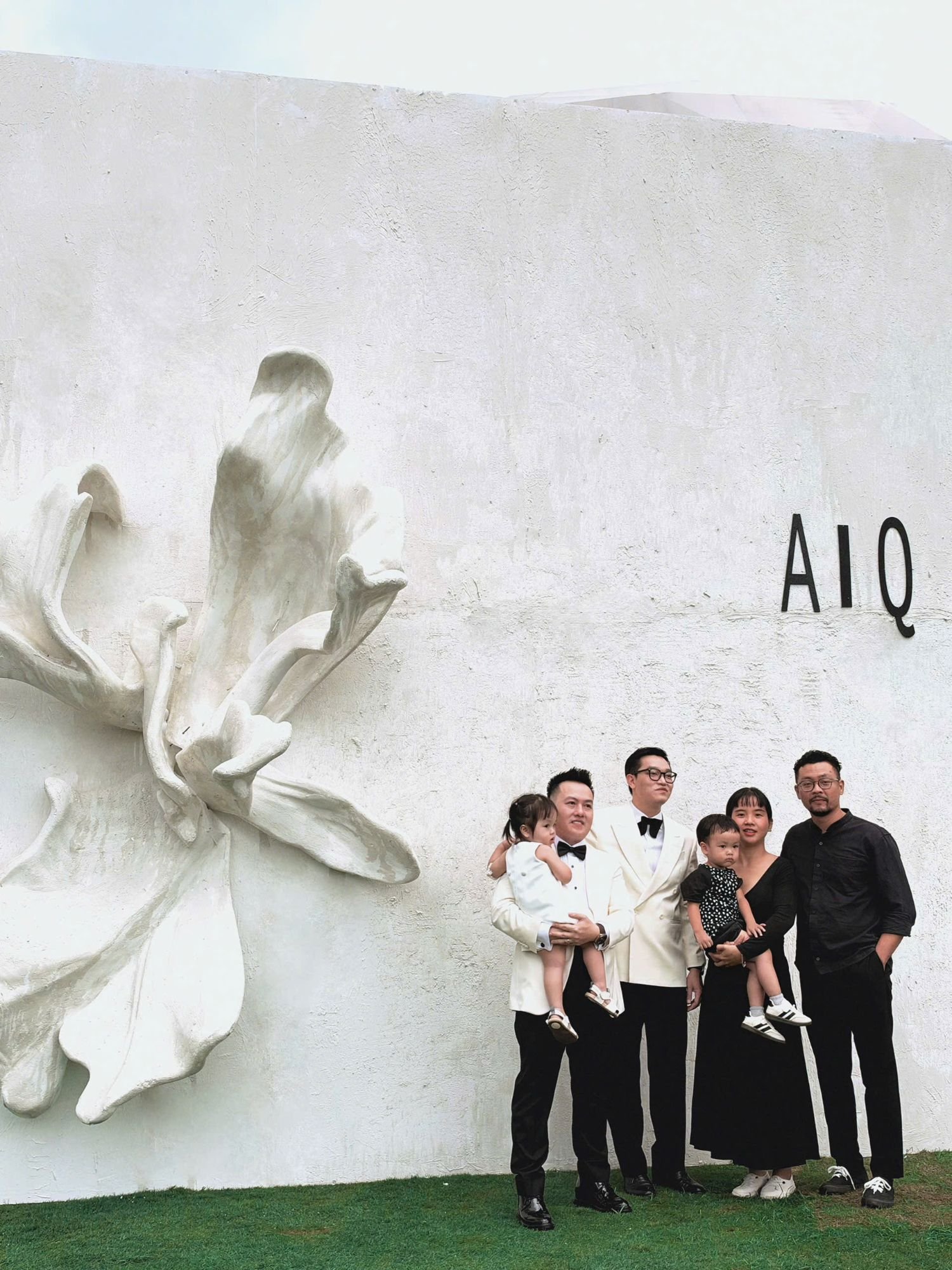 Hai chú rể cùng gia đình trước tường hoa điêu khắc và chữ A | Q